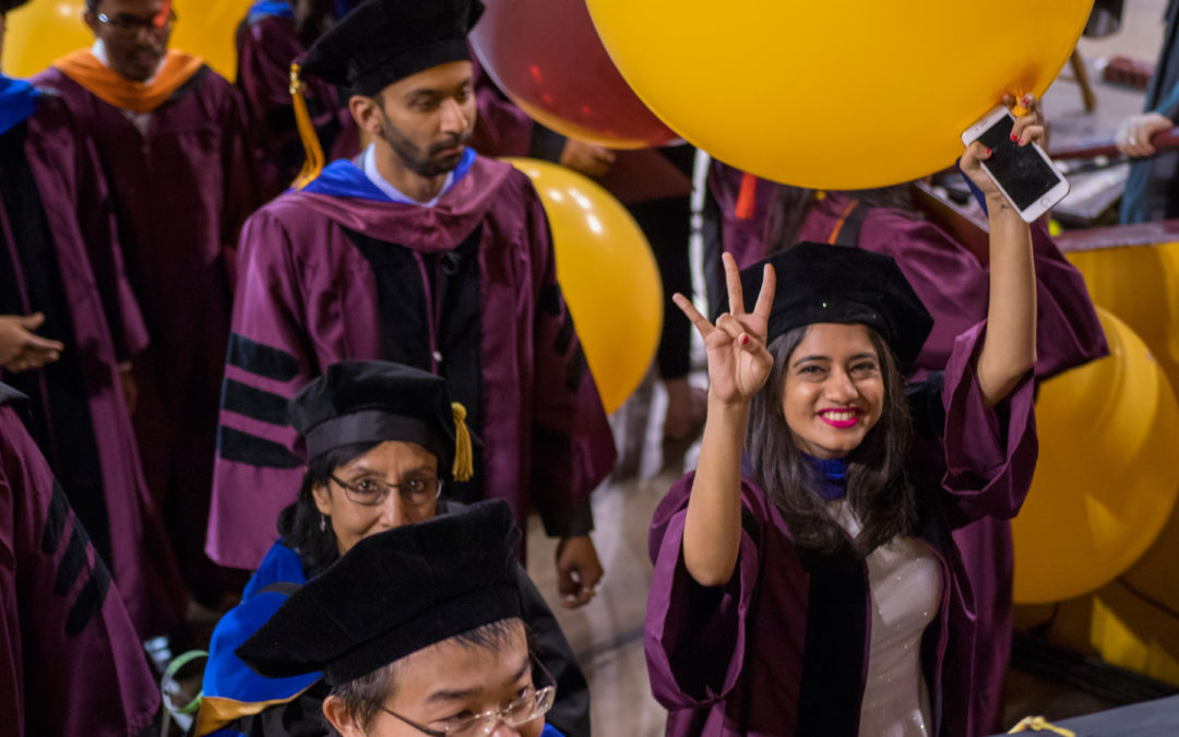 A student holds a balloon in one hand and holds an ASU pitchfork gesture in the other, as she smiles leaving Convocation