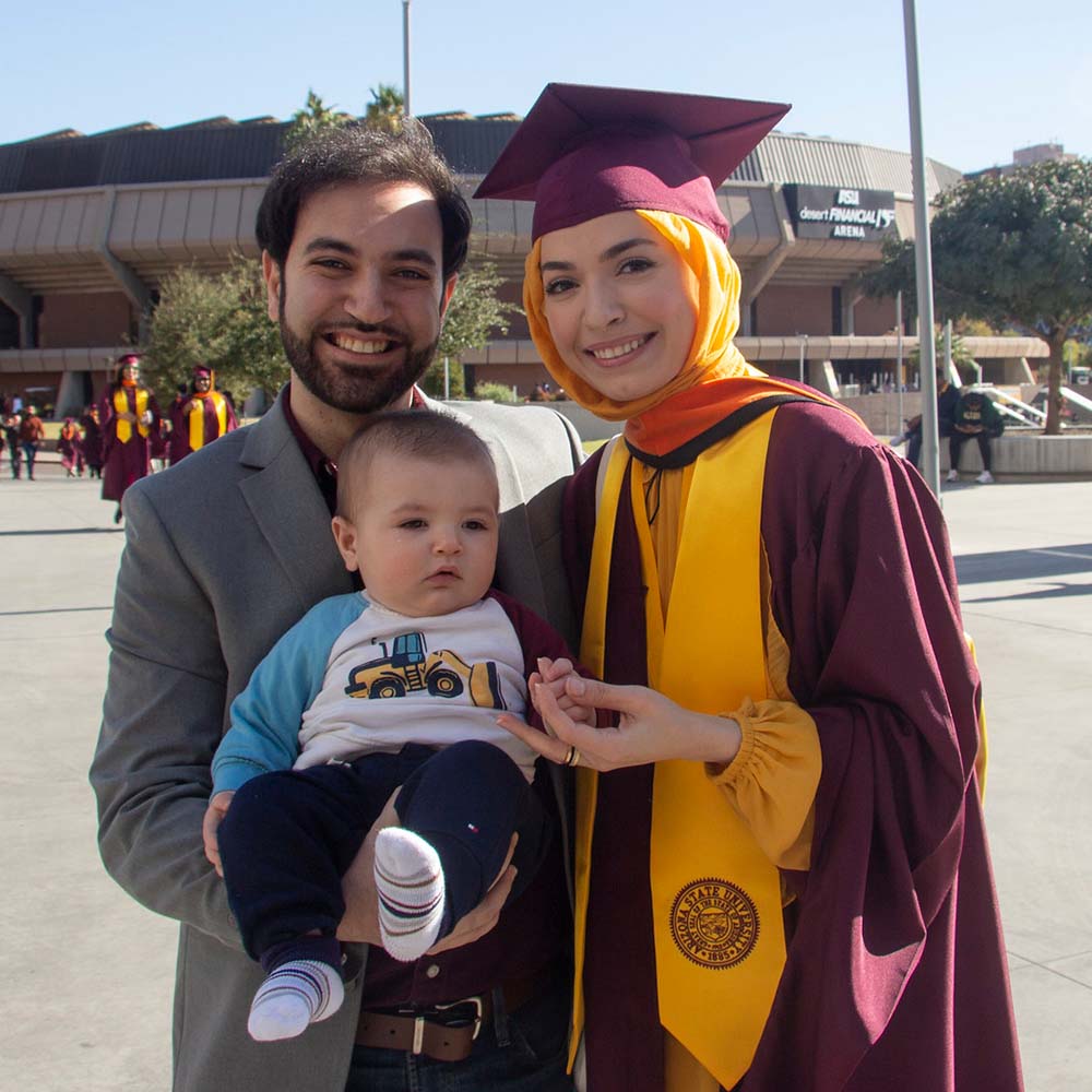 A graduate and her husband stand together holding their baby outside Desert Financial Arena.