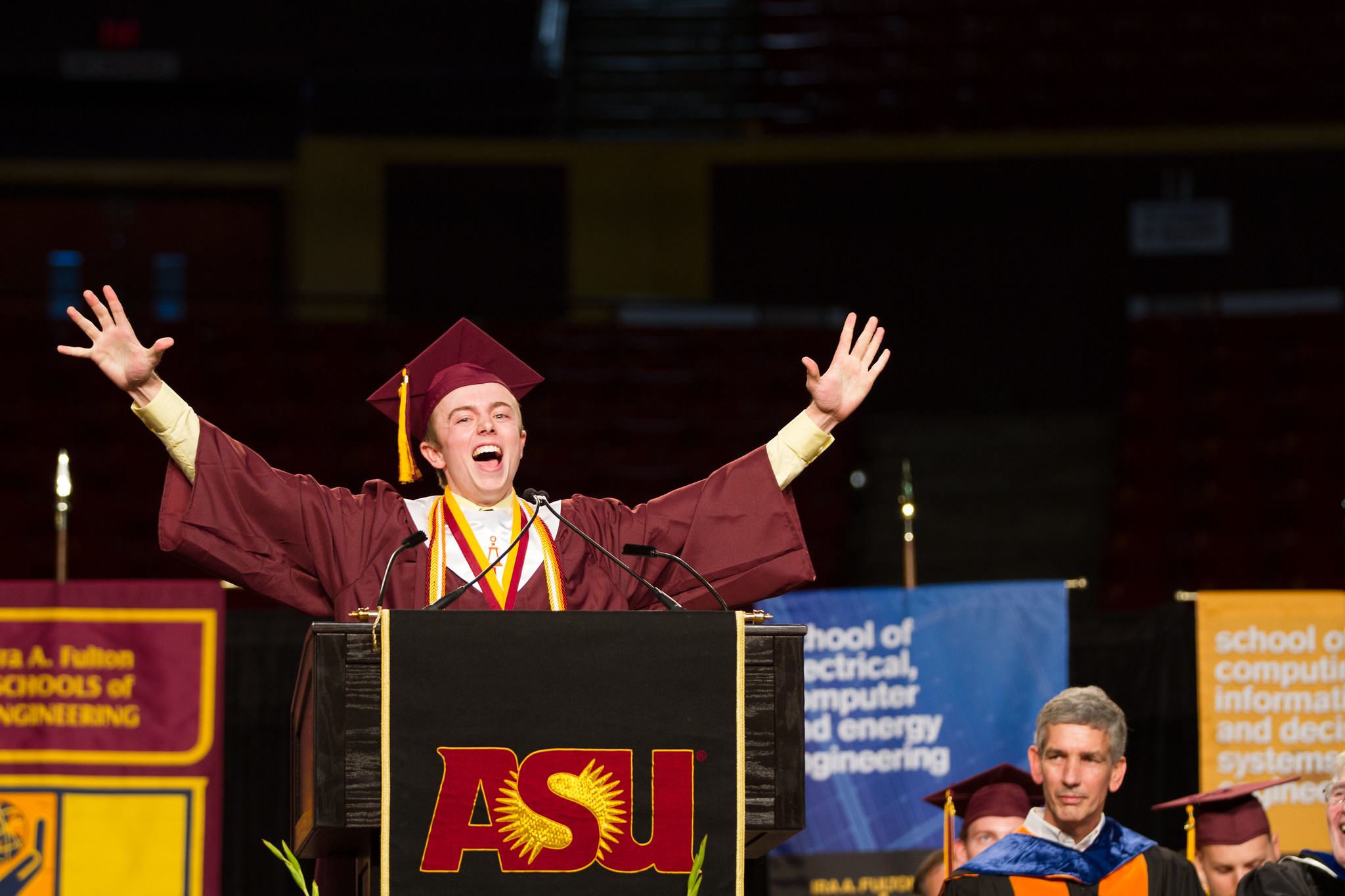 A graduate stands at the convocation podium with his arms held high in celebration