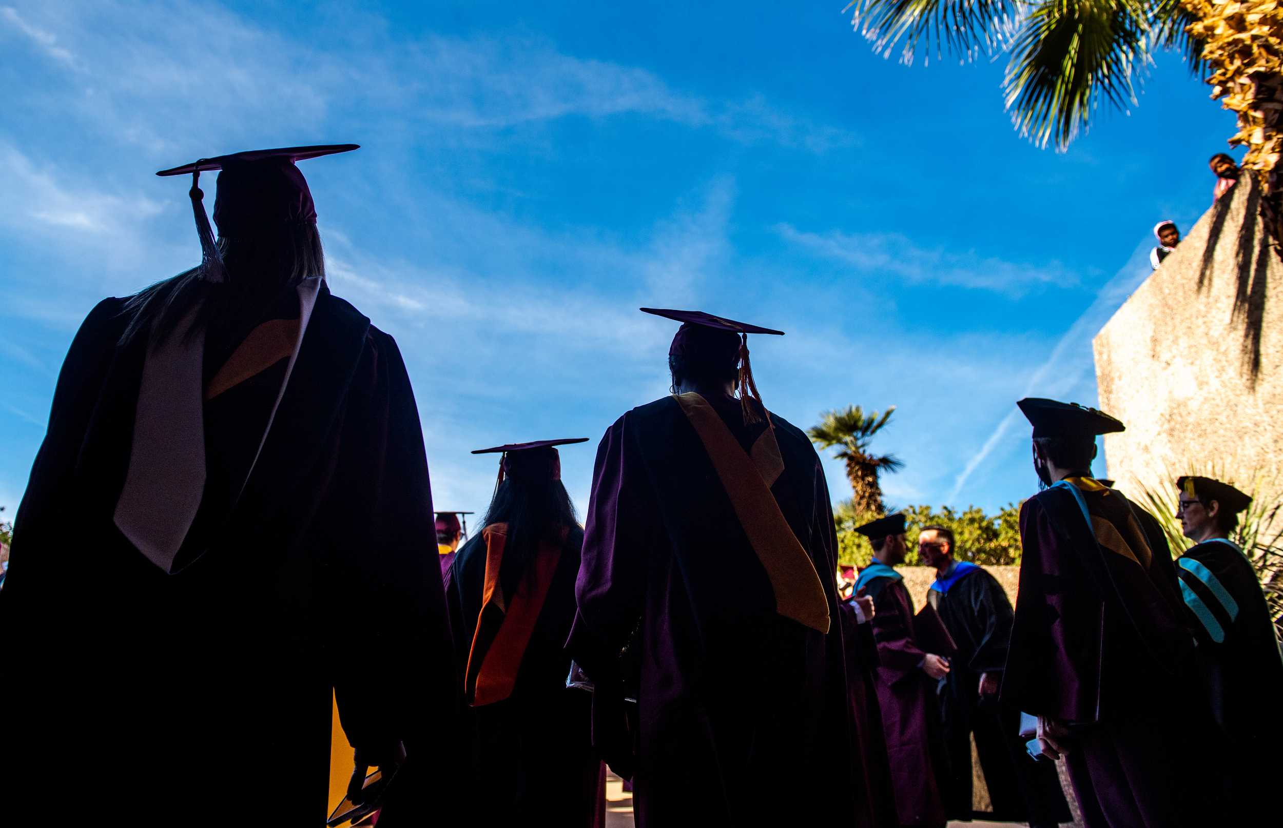 A scene from behind of ASU graduates walking outside in the bright sunshine towards the ASU graduate commencement ceremony.