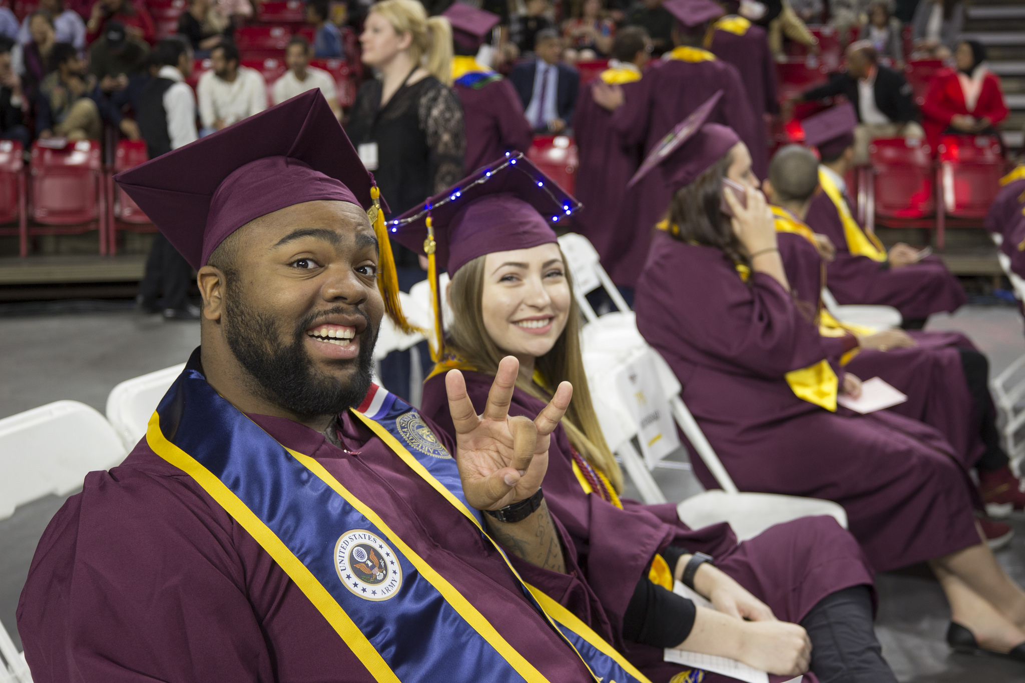 This image shows two smiling graduates sitting together at their convocation ceremony. The graduate in the foreground is wearing a U.S. Army sash and proudly displaying the ASU "pitchfork" hand sign, while the other graduate, wearing a decorated cap with lights, smiles for the camera.