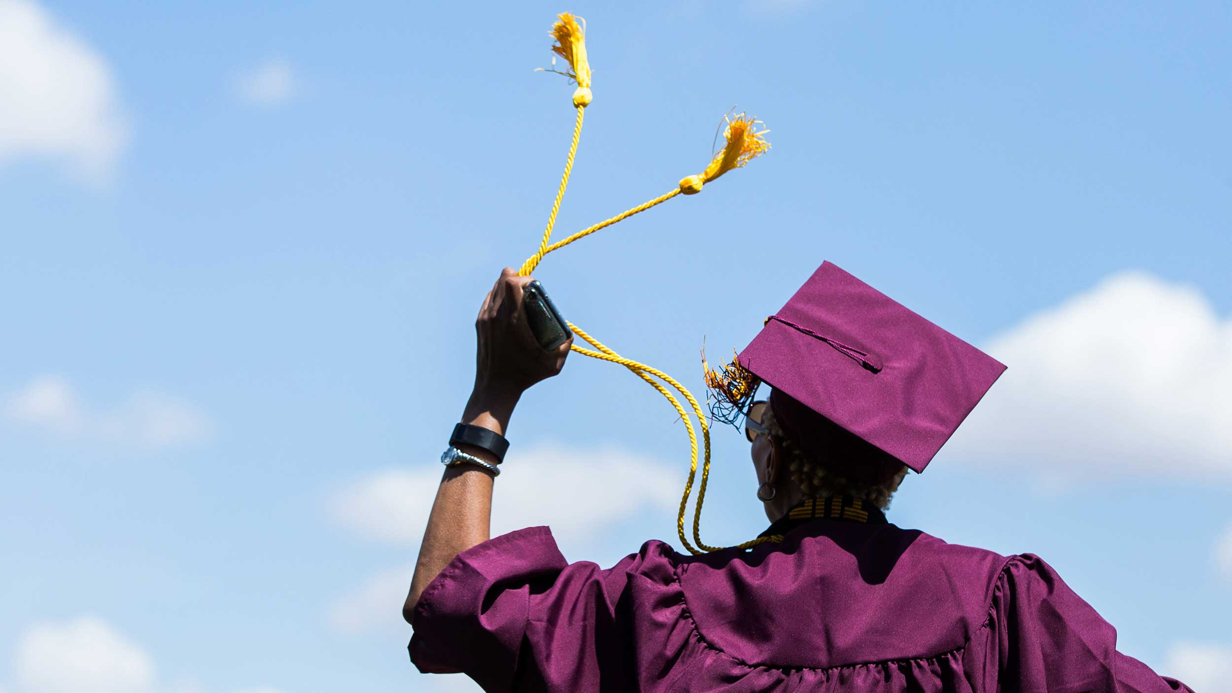 An ASU grad is shown jumping in his cap and gown against a bright blue sky