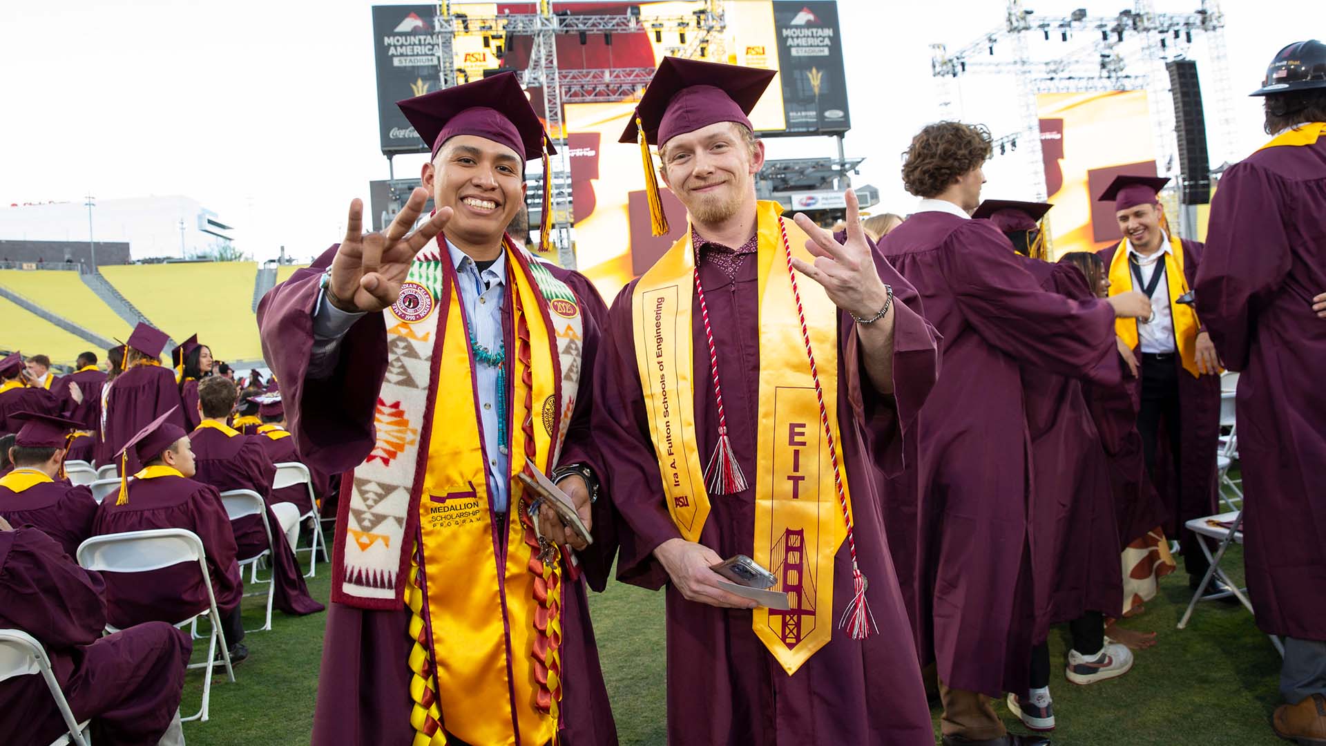 Two male graduates, dressed in ASU regalia, stand together smiling at the Spring 2025 Ira A. Fulton Schools of Engineering Undergraduate Convocation ceremony. They hold their hand in the ASU pitchfork gesture of school spirit. Both wear stoles representing special honors.