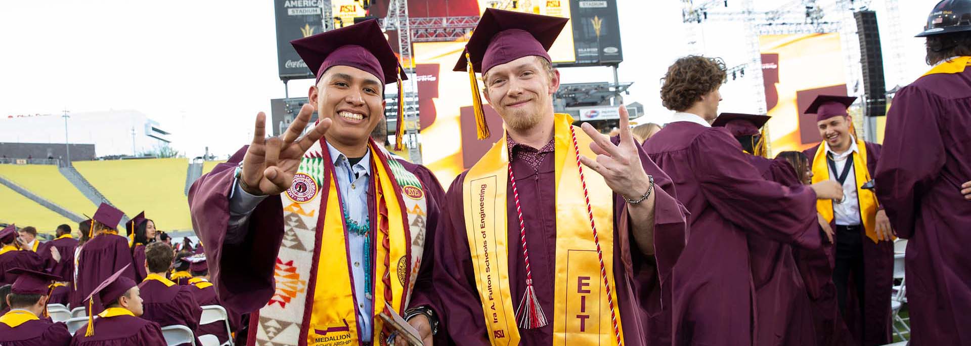 Two male graduates, dressed in ASU regalia, stand together smiling at the Spring 2025 Ira A. Fulton Schools of Engineering Undergraduate Convocation ceremony. They hold their hand in the ASU pitchfork gesture of school spirit. Both wear stoles representing special honors.