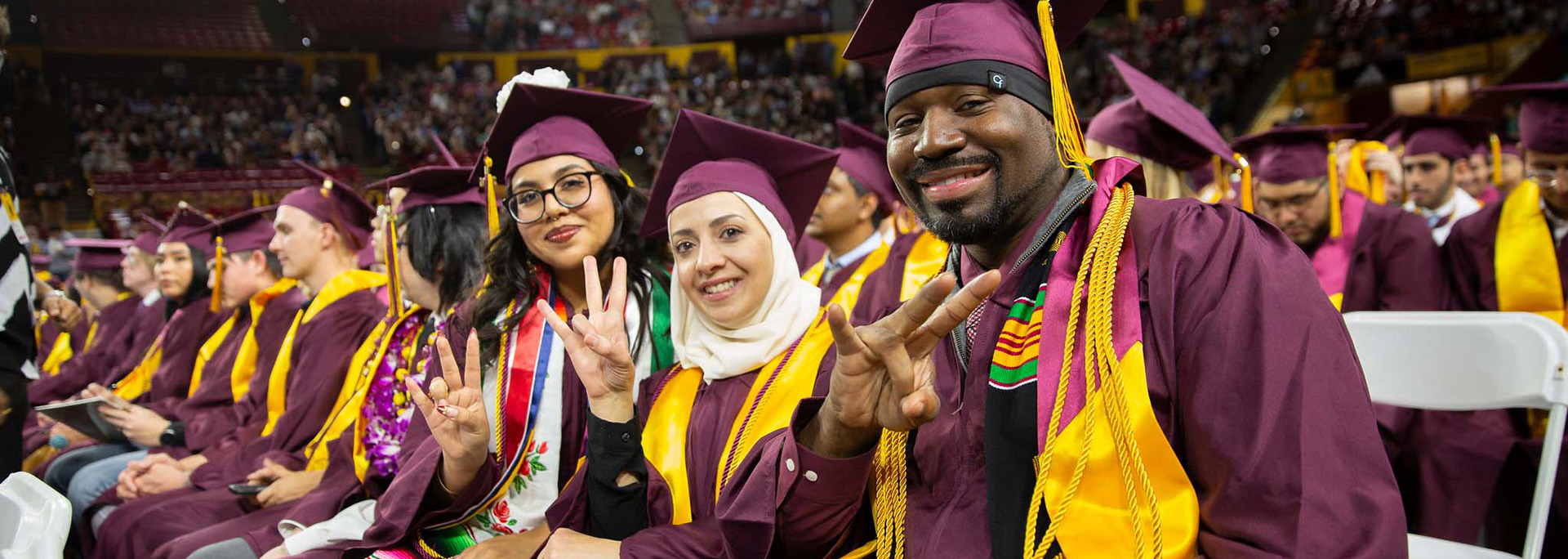 Three graduates in maroon caps and gowns with gold stoles sitting in a row of other graduates, smile and pose for the camera, each holding up the university's pitchfork hand sign.