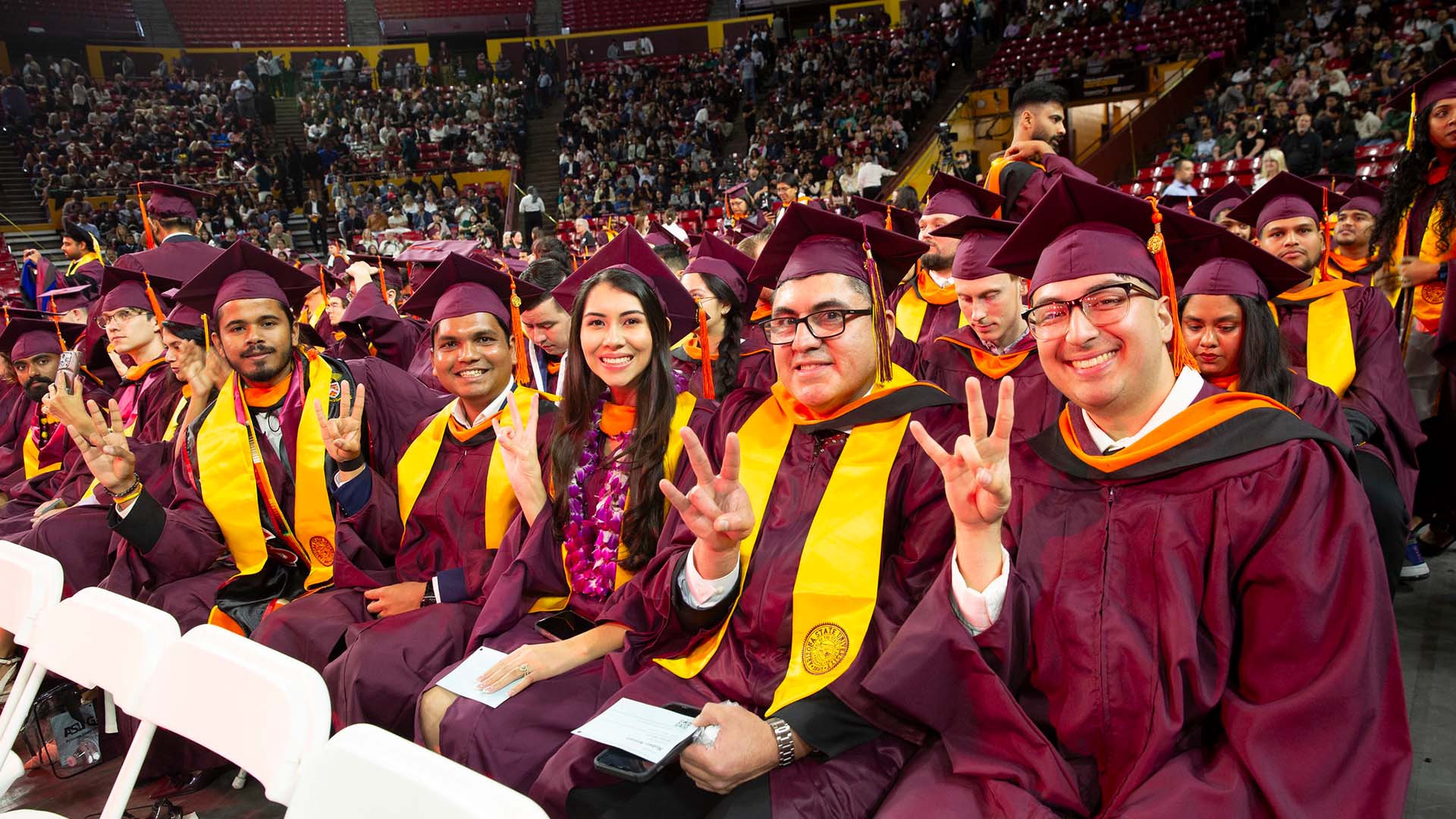 A group of graduates in maroon caps and gowns sit together at a ceremony, smiling and holding up pitchfork signs, with a large audience behind them.