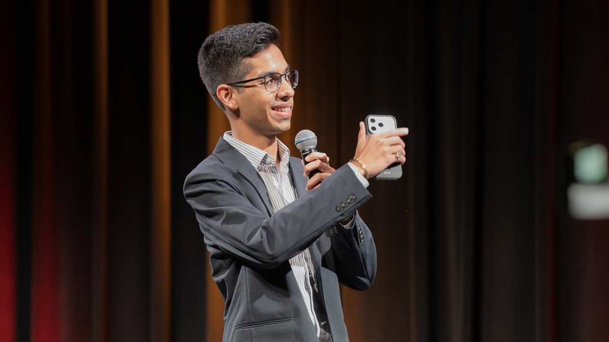 Vijay Abhirup stands on a stage wearing a gray suit, holding a microphone in one hand and his phone in the other, speaking with the audience in front of a maroon stage curtain.