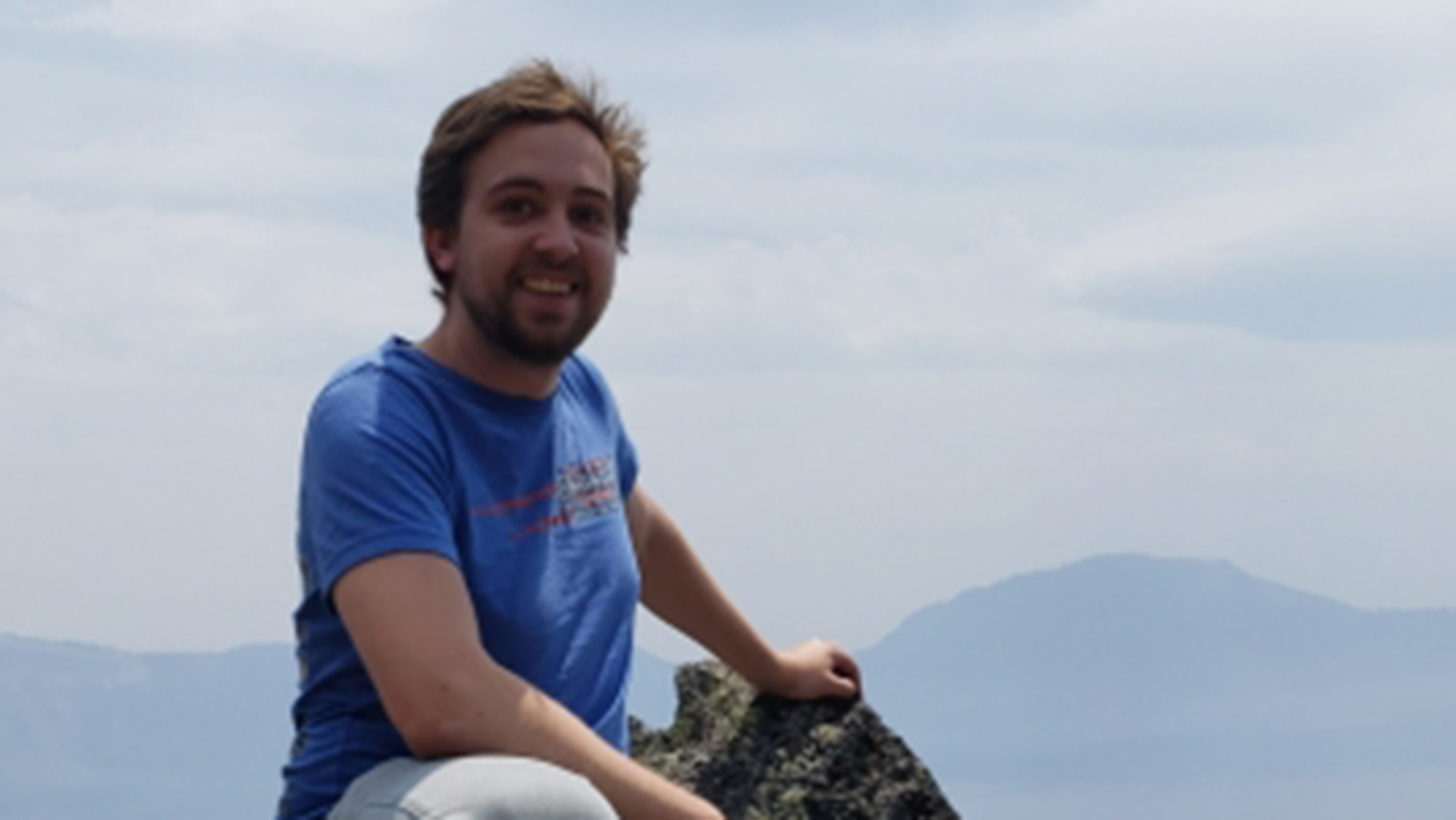 Man in a blue shirt sits on a rock with mountains in the background.