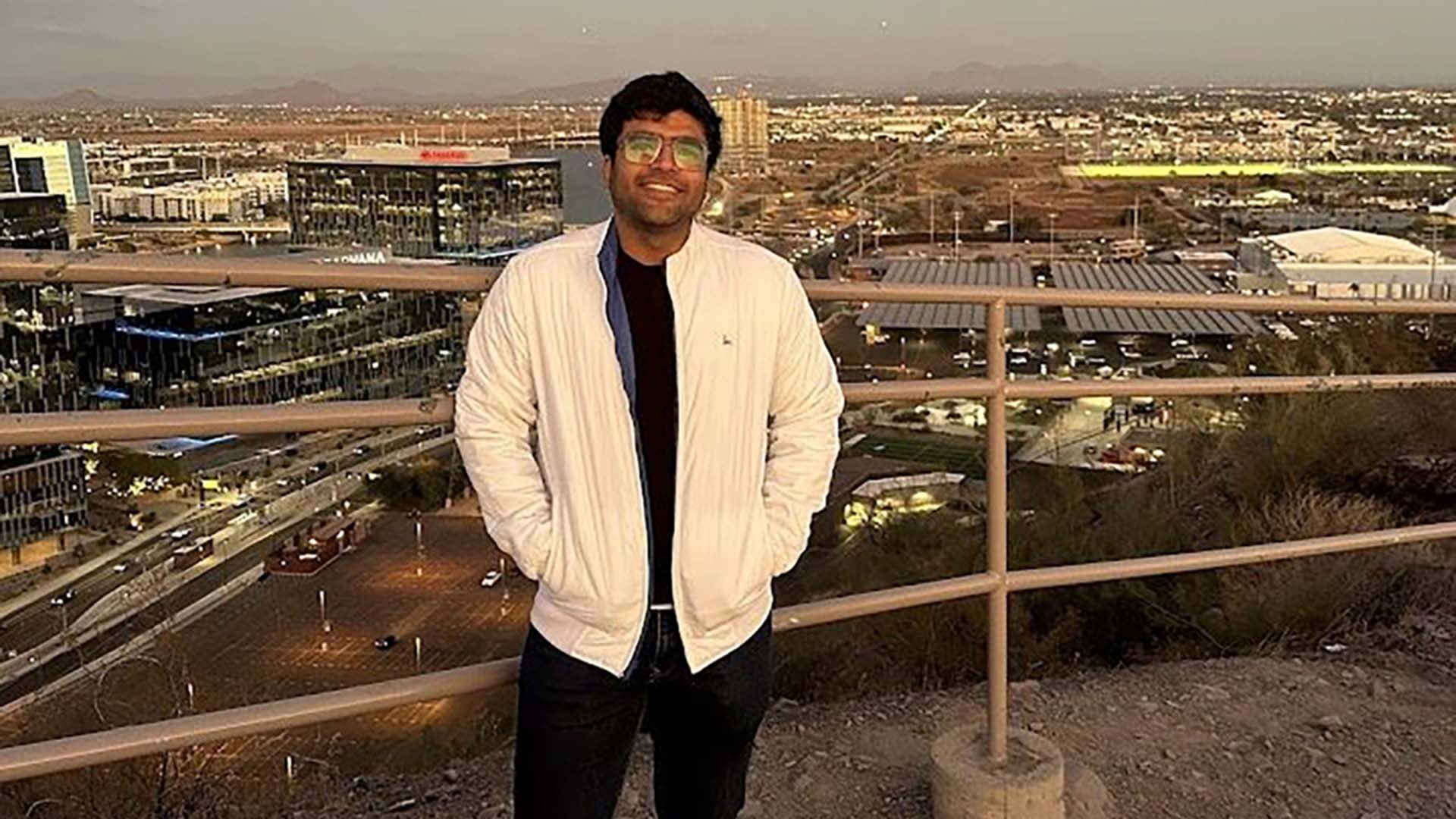 Man wearing glasses and a white jacket stands on a hill overlooking a city at sunset.