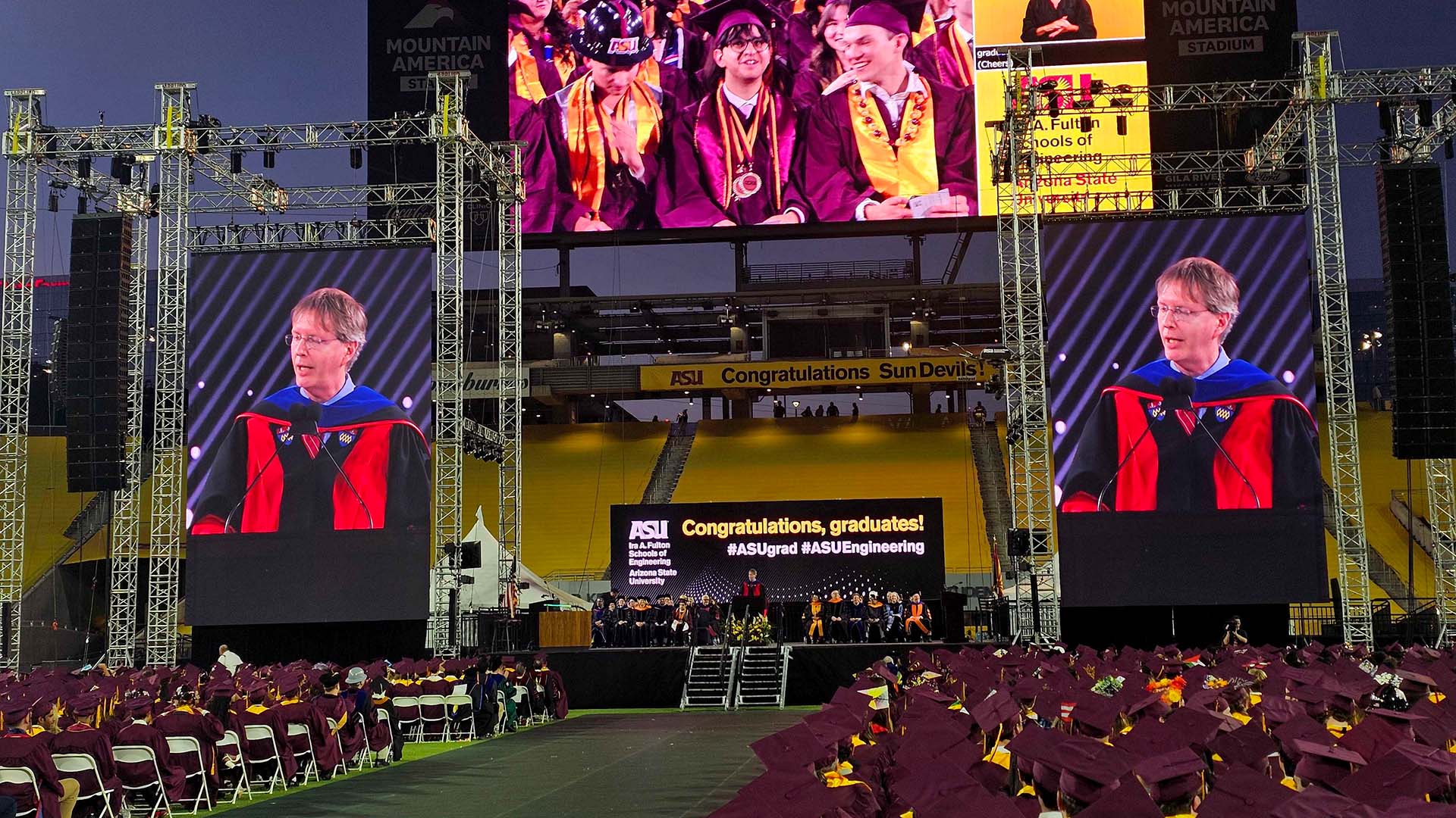 Wide view of the ASU Fulton Schools Convocation ceremony inside Mountain America Stadium. Graduates in maroon caps and gowns sit facing a stage where a speaker, Dean Kyle Squires, stands at a podium. Large vertical screens on both sides display a close-up of Squires, and additional screens above show graduates in the audience.