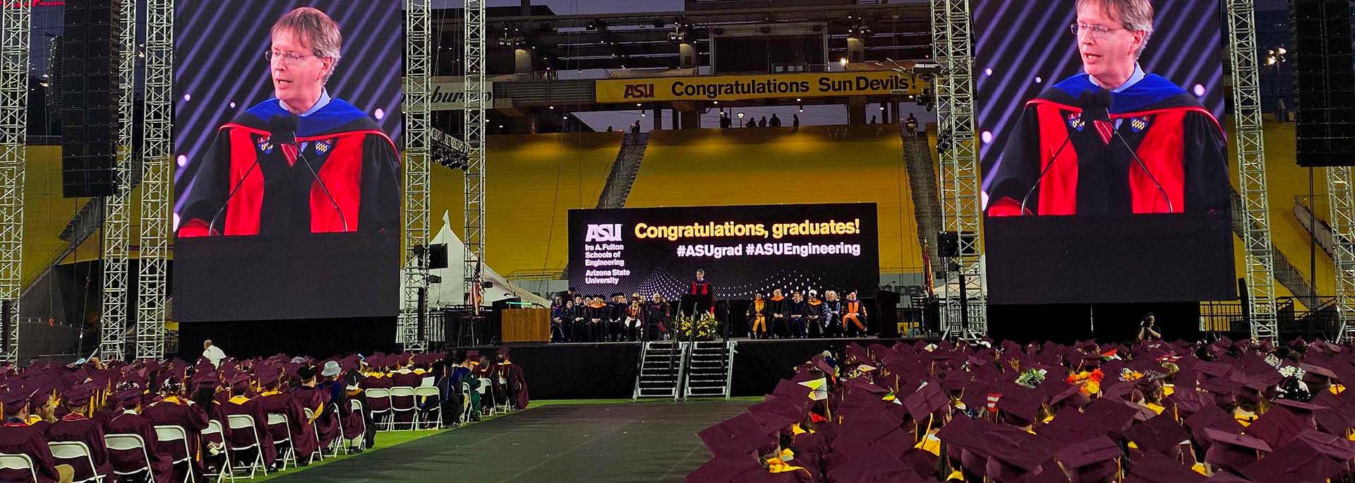 Wide view of the ASU Fulton Schools Convocation ceremony inside Mountain America Stadium. Graduates in maroon caps and gowns sit facing a stage where a speaker, Dean Kyle Squires, stands at a podium. Large vertical screens on both sides display a close-up of Squires.